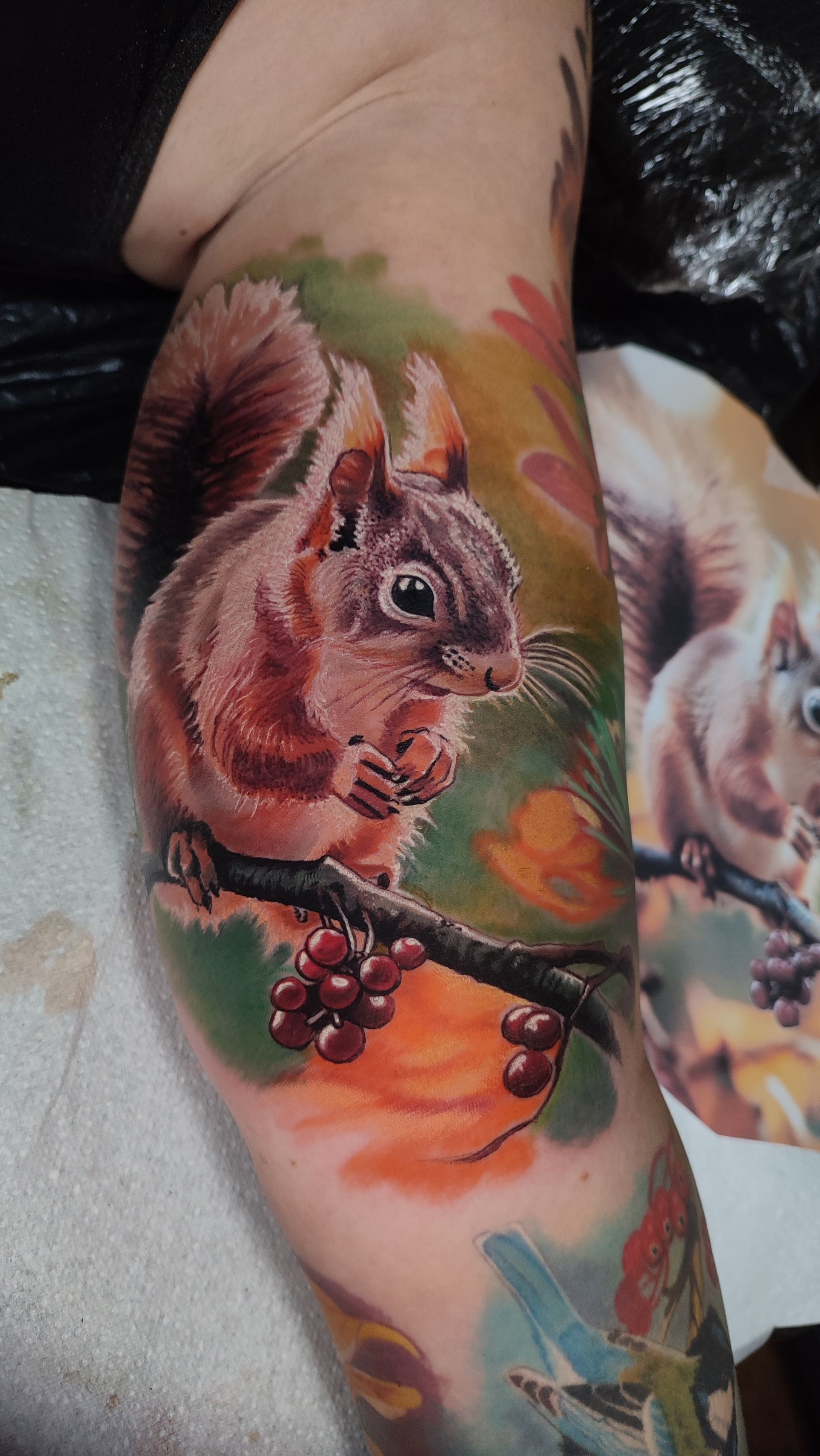 Tattoo of a squirrel perched on a branch with red berries, surrounded by colorful leaves and a blurred background.