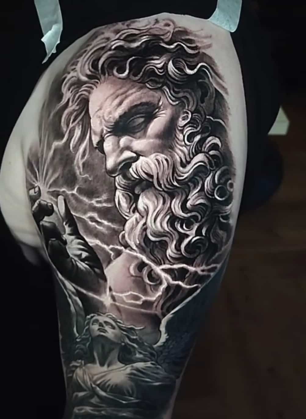 Tattoo of Zeus with lightning, holding a glowing object. Dark tones, detailed face, and a figure below.