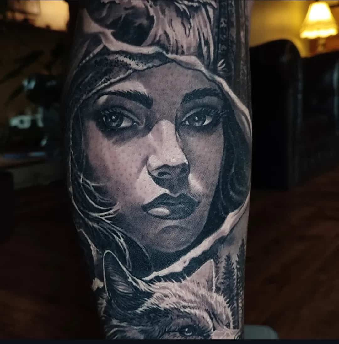 Black and white tattoo of a woman's face surrounded by a wolf and forest.