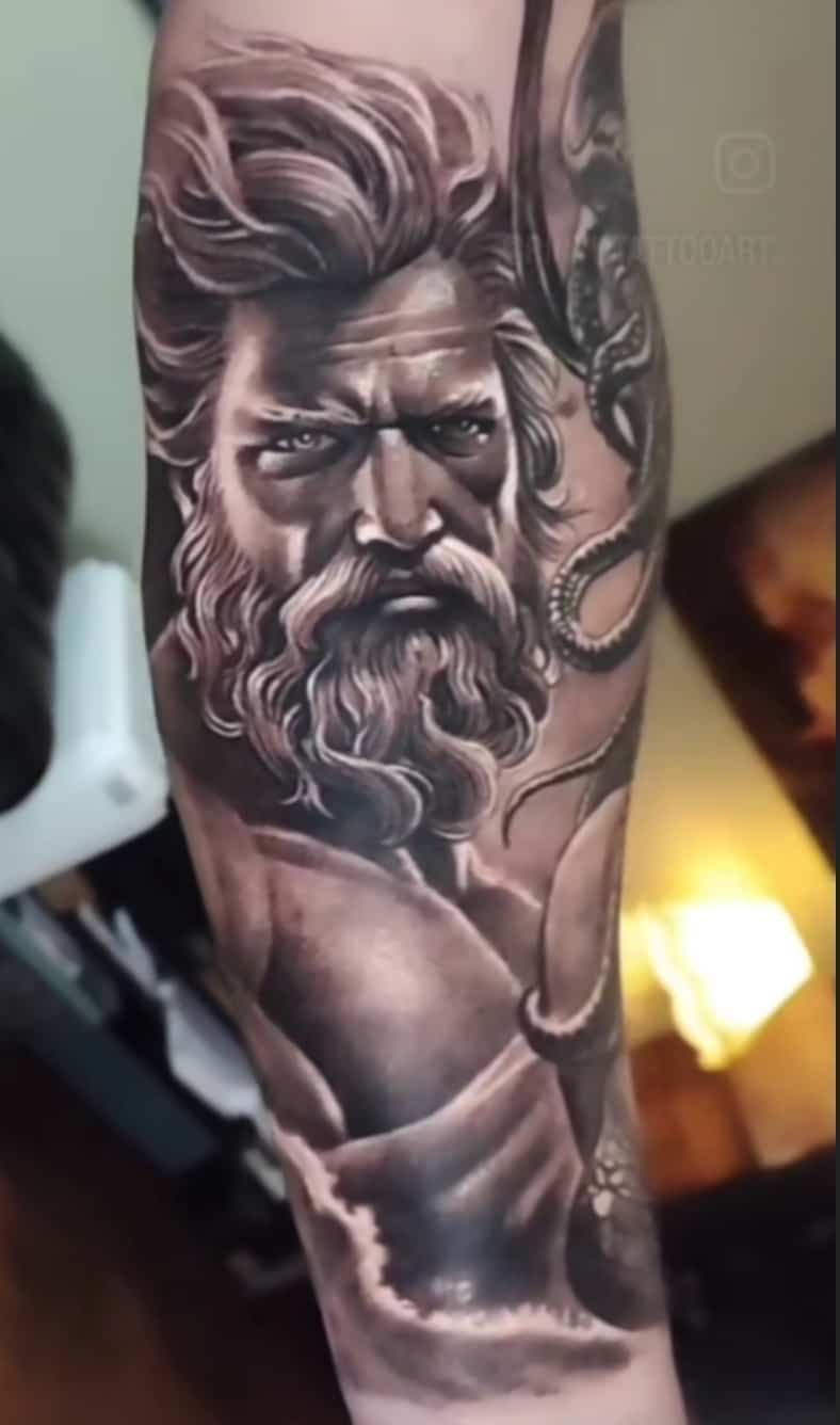 Black and white tattoo of a stern-faced man with a beard, in a classical pose, on an arm.