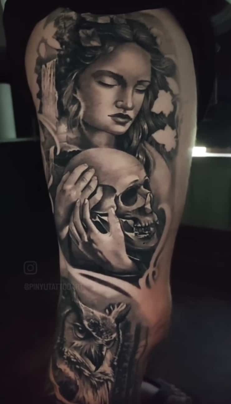Tattoo of a woman with closed eyes holding a skull, surrounded by flowers and a wolf. Black and gray.