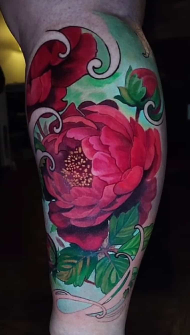 Vibrant peony tattoo on a leg. Red petals with green leaves and light green background.