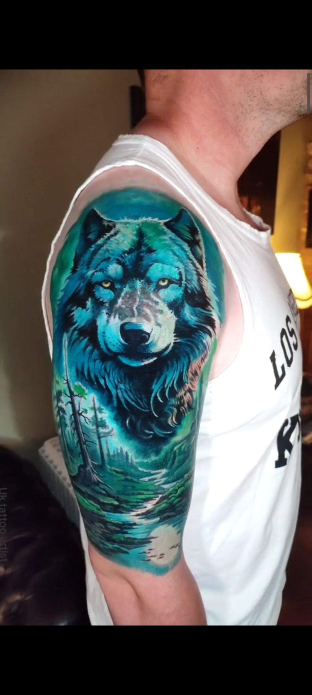 A blue and green wolf tattoo on a man's arm, depicting a forest scene.
