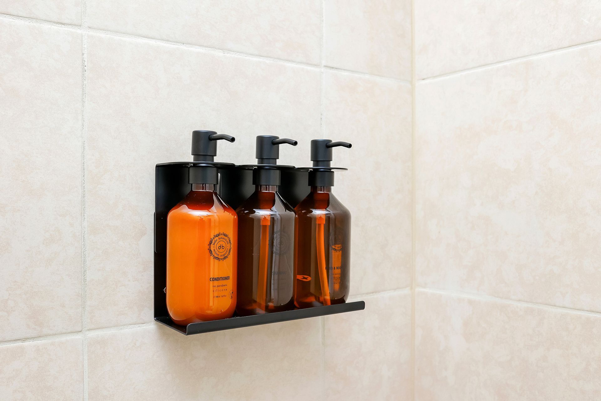 Black wall-mounted dispenser rack holding three amber-colored bottles with black pumps on a tiled wall.