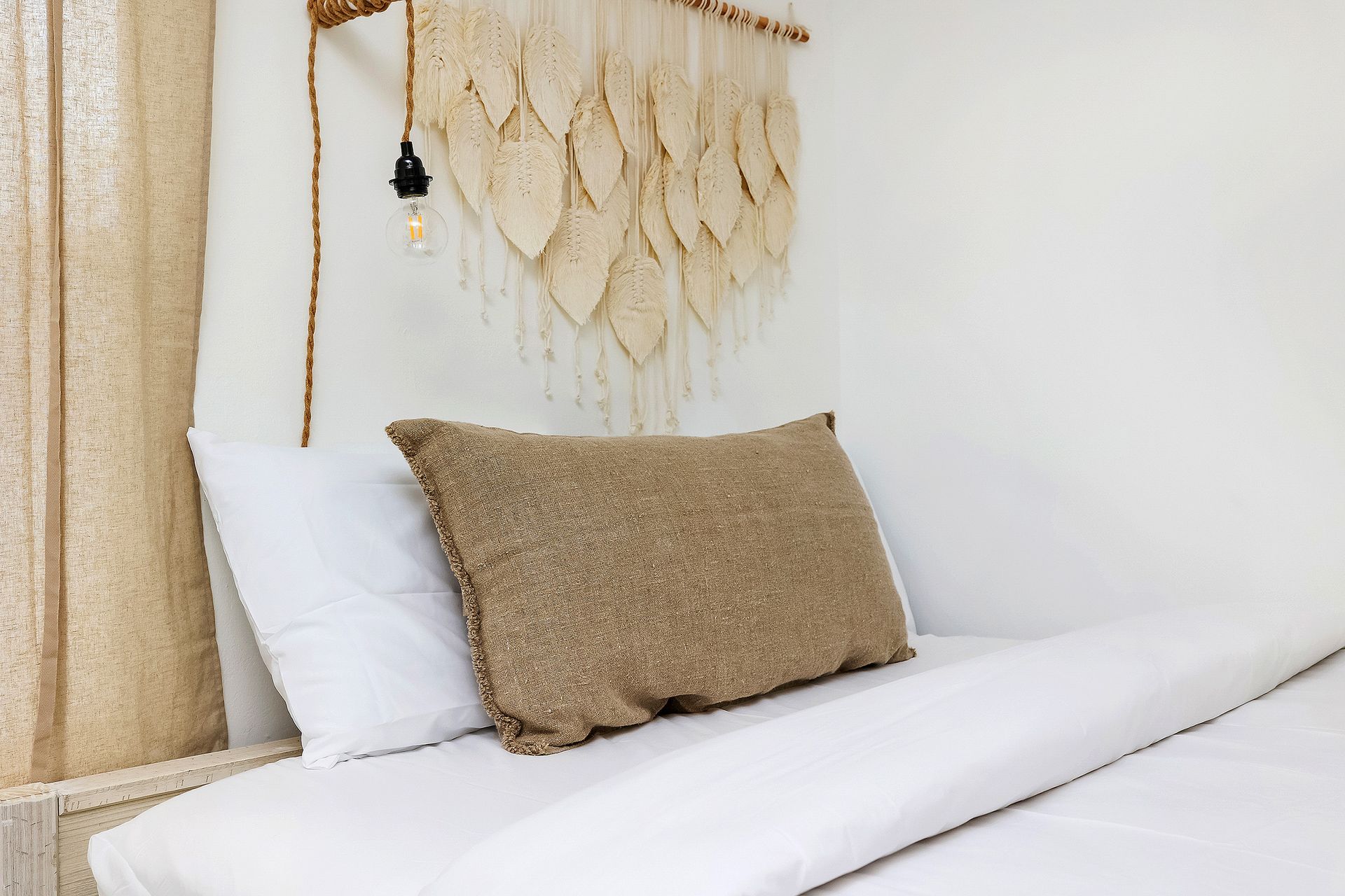 Bed with white linens, beige pillow, and wall hanging in a bright room.