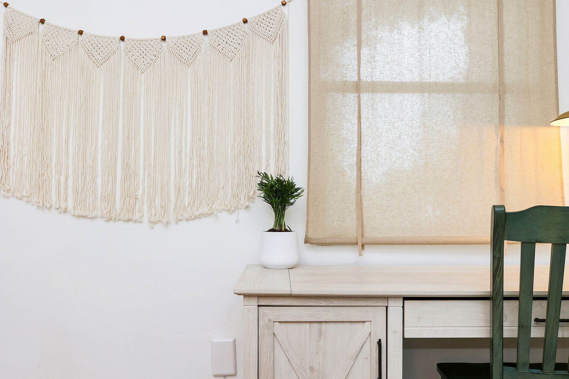 Beige macrame wall hanging above a desk with a plant and window with beige blinds.