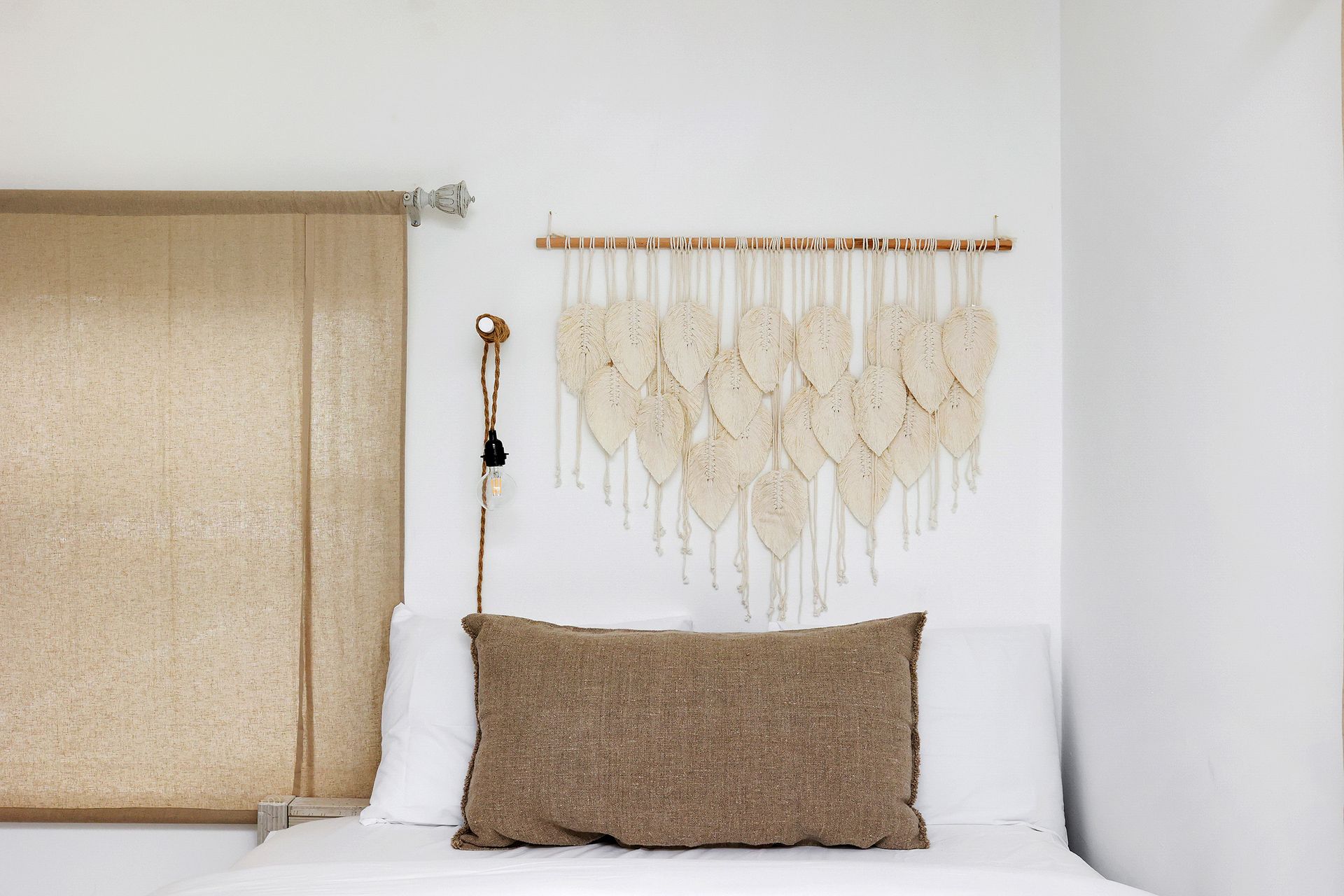 Bedroom with macrame wall hanging above a bed with tan pillow and window with beige blind.
