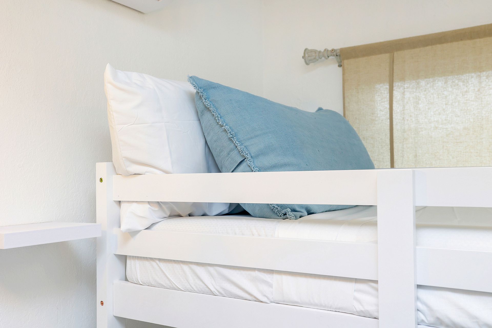 White bunk bed with white bedding, a white pillow, and a blue pillow. Window in the background.