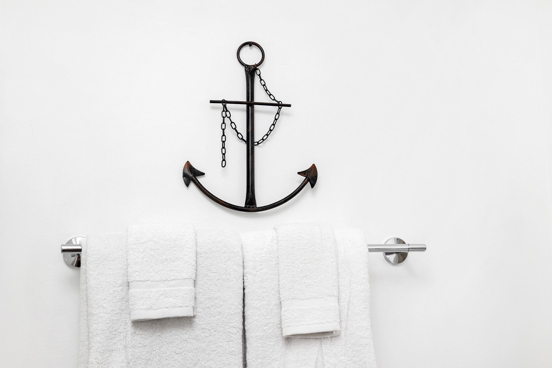 Anchor wall decor above a chrome towel bar with white towels against a white wall.