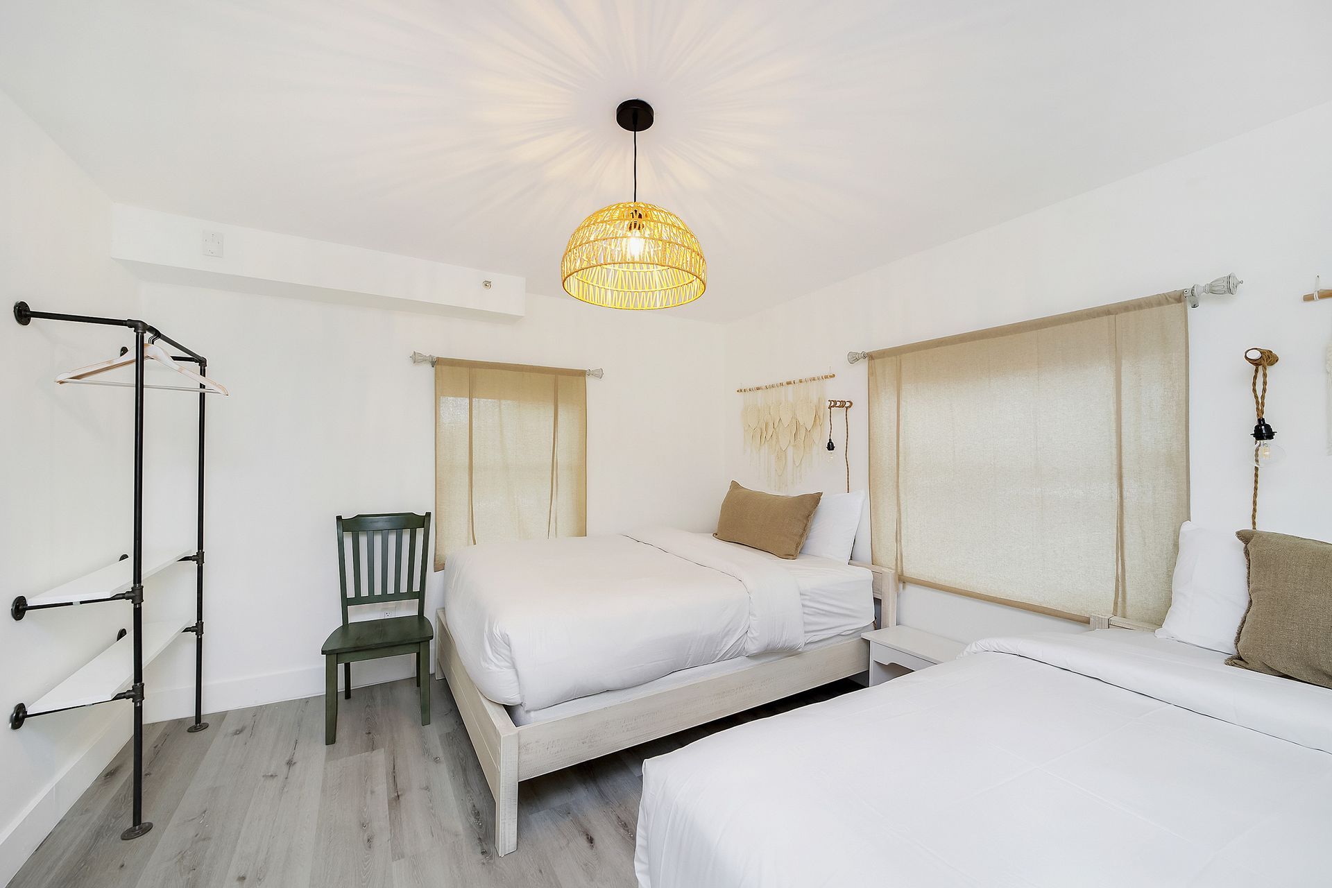 Bedroom with two beds, white walls, gray wood floor, woven light fixture, and a green chair.