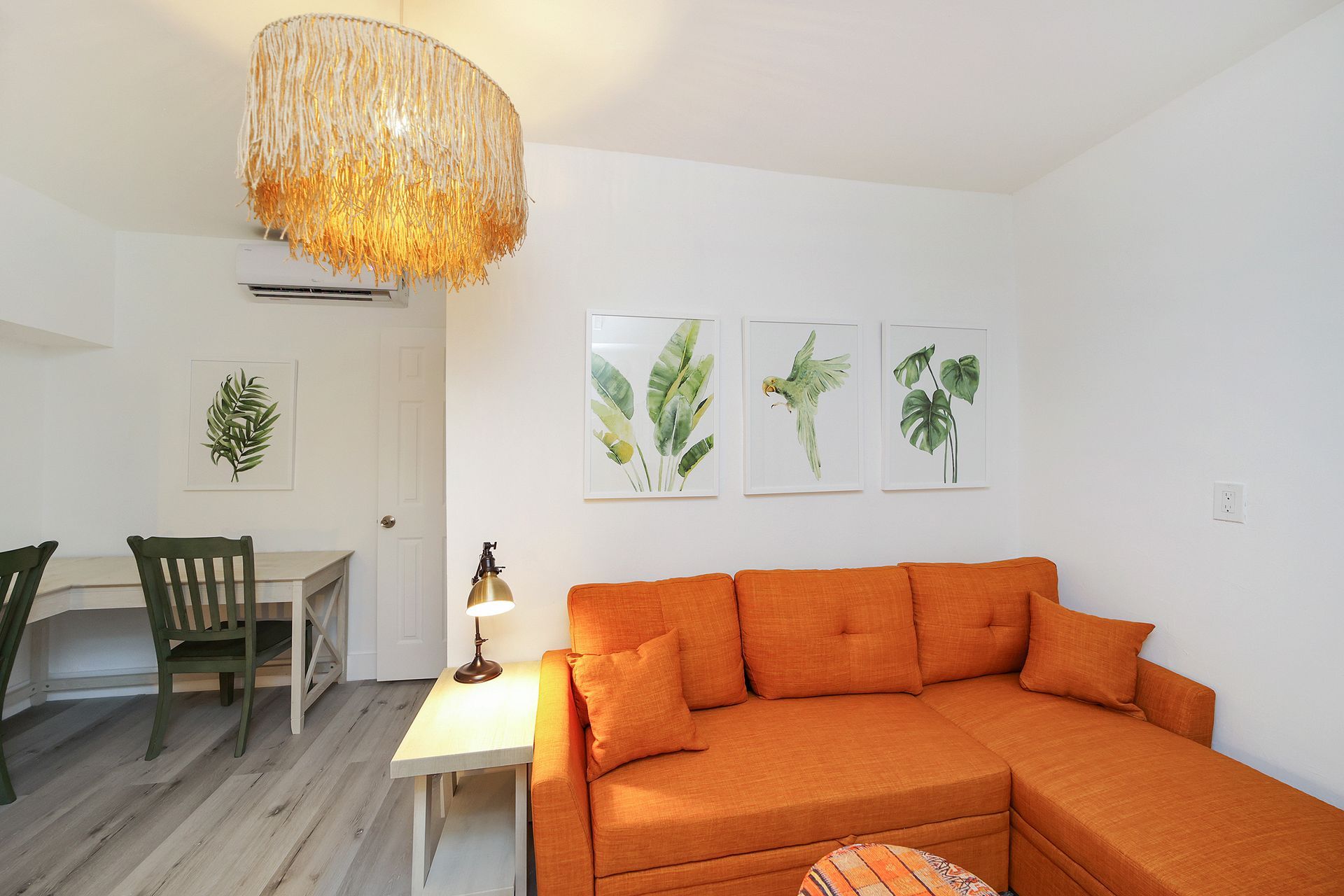 Bright orange sofa, art, and light fixture in a white-walled room. Wooden desk with chair to the left.