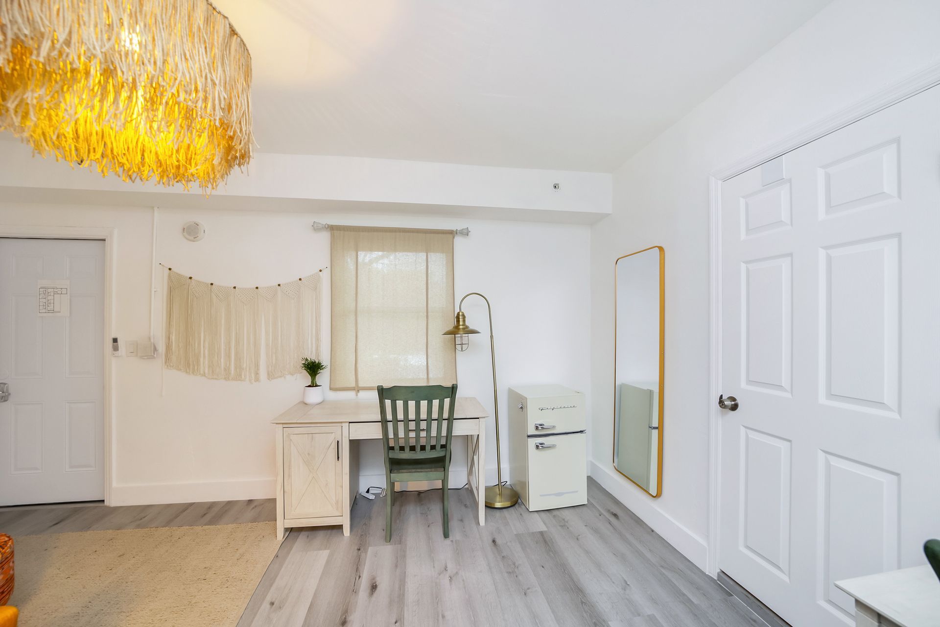 Bright white room with desk, chair, mini-fridge, mirror, and two doors. Light fixture hangs from the ceiling.