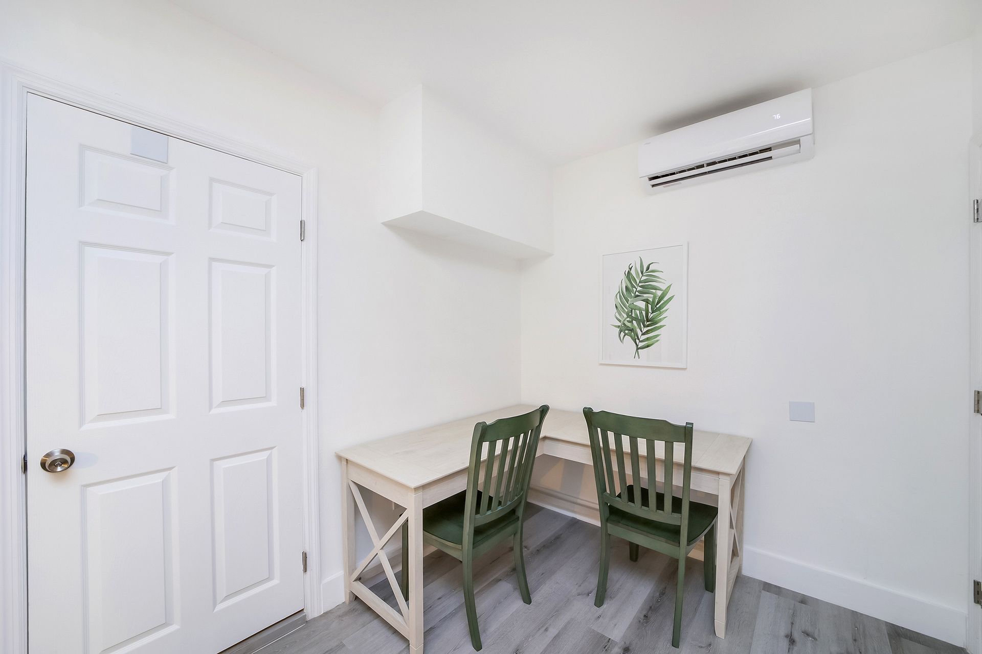 White room with desk, two green chairs, door, air conditioner, and art.