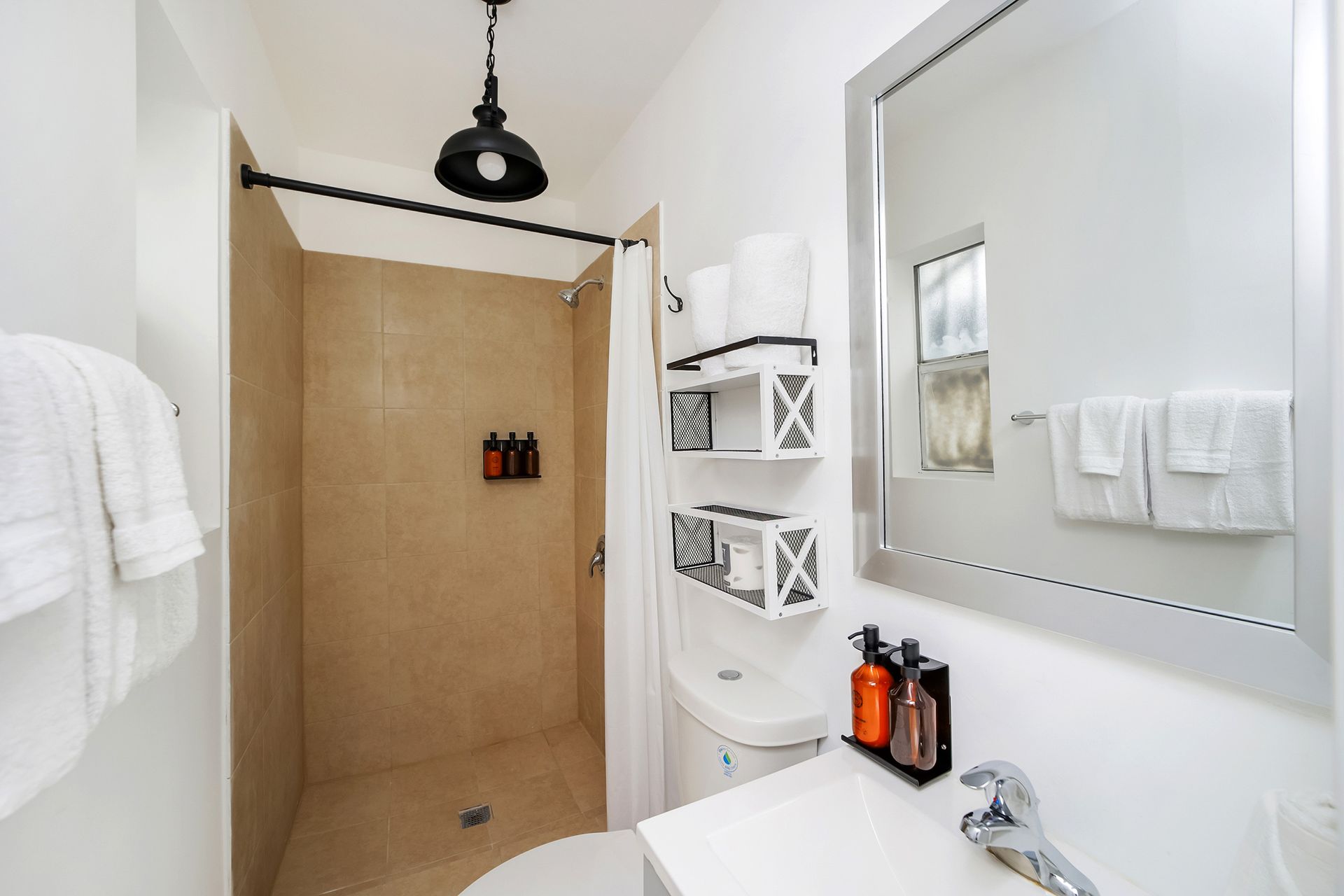 Small white bathroom with shower, toilet, and sink. Towels and shelving are present. Black light fixture.