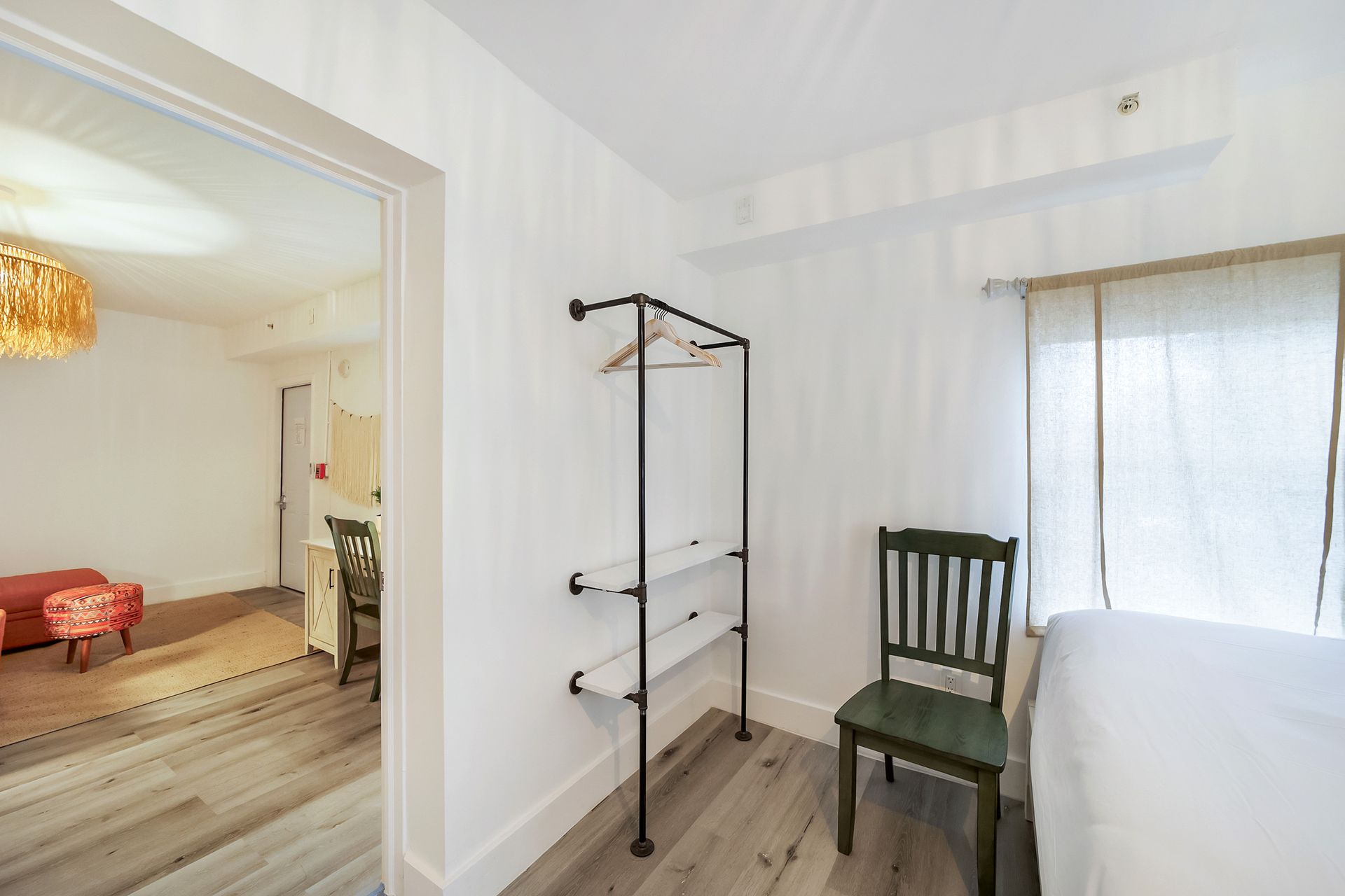 Bedroom interior with white walls, a clothing rack, green chair, and a view into another room.