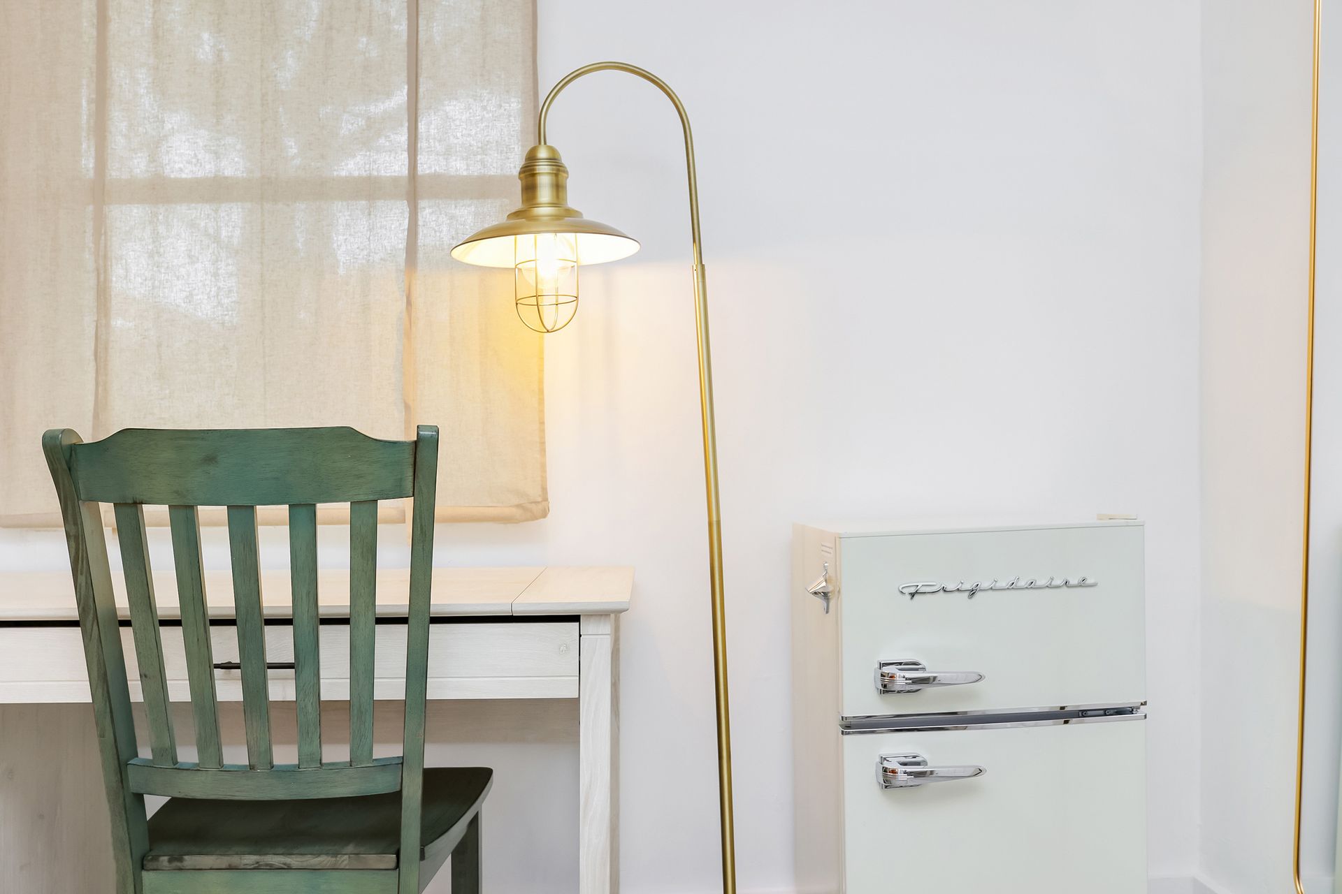 Green chair at white desk, arched brass lamp, vintage refrigerator in a white room.