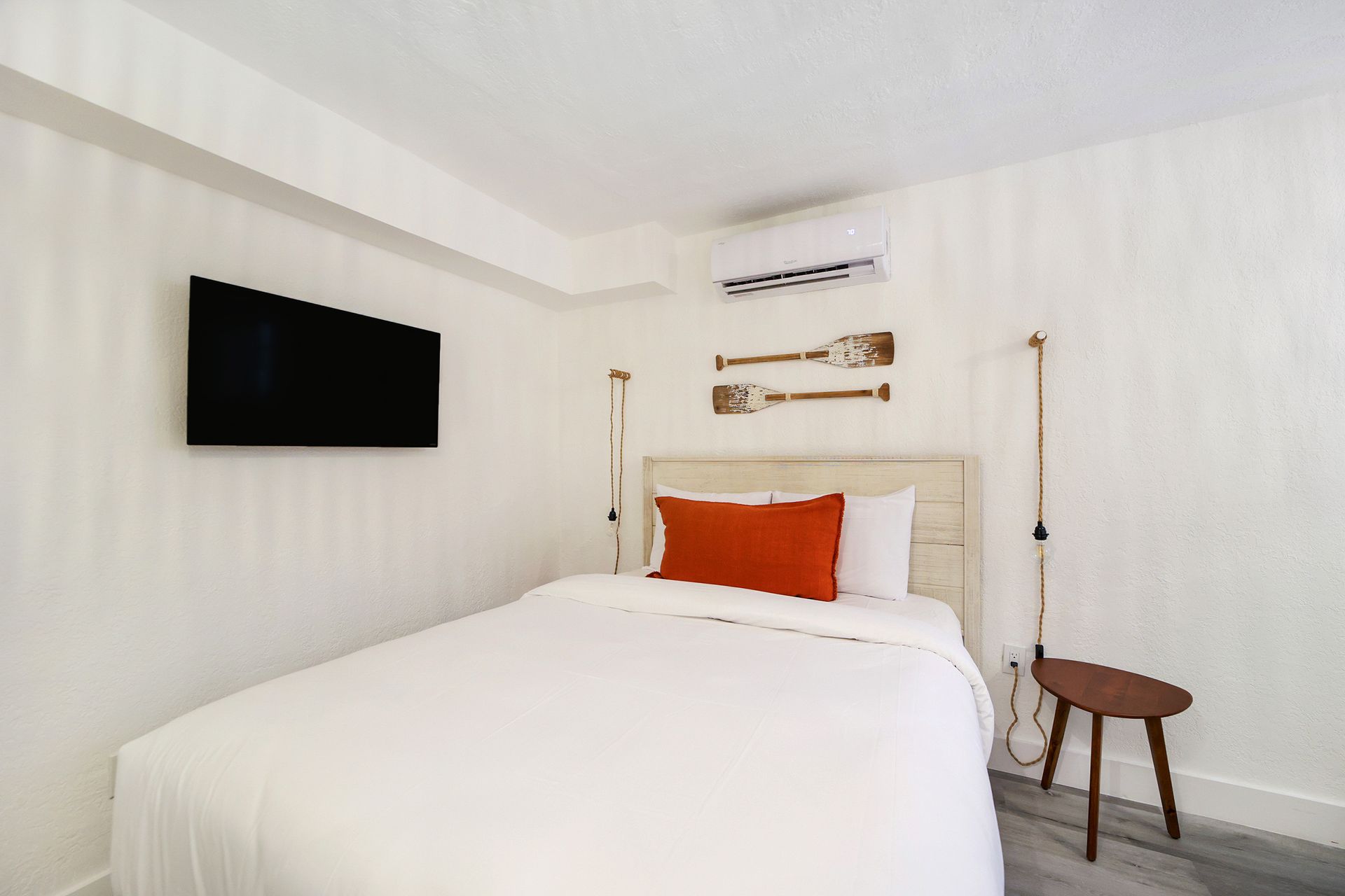 White bedroom with bed, TV, air conditioner, wooden oar decor, and small stool.