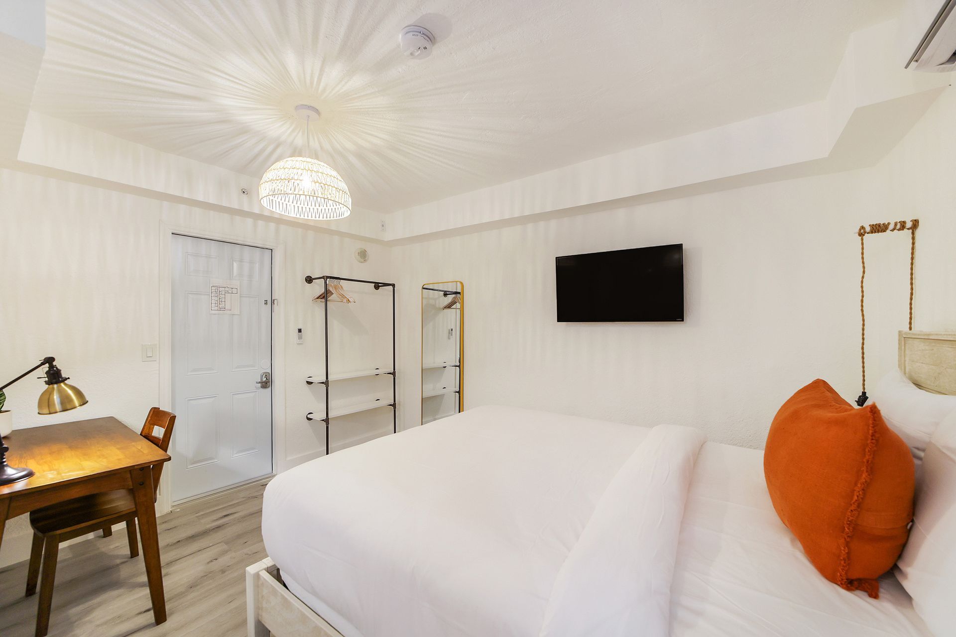 White bedroom with bed, desk, TV, hanging clothes rack, and shower. Orange pillow on bed.