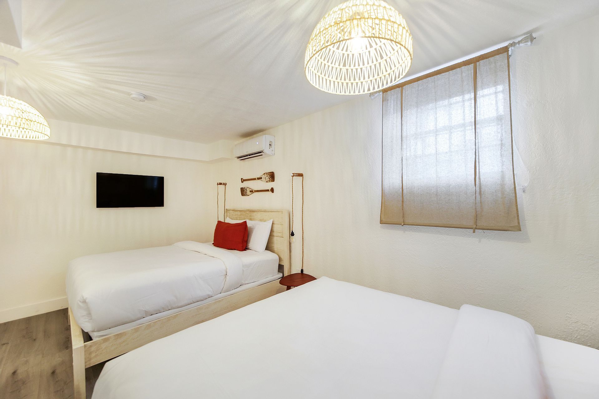 Bright, white bedroom with two beds, red pillow, TV, and woven pendant lights.