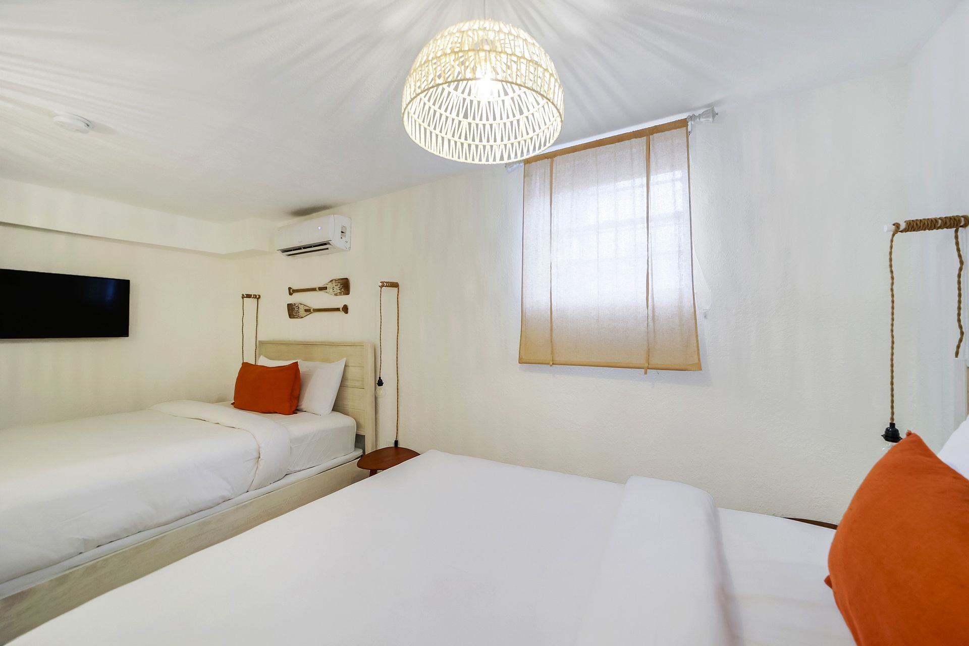 Bright hotel room with two beds, orange pillows, window, and wicker light fixture.
