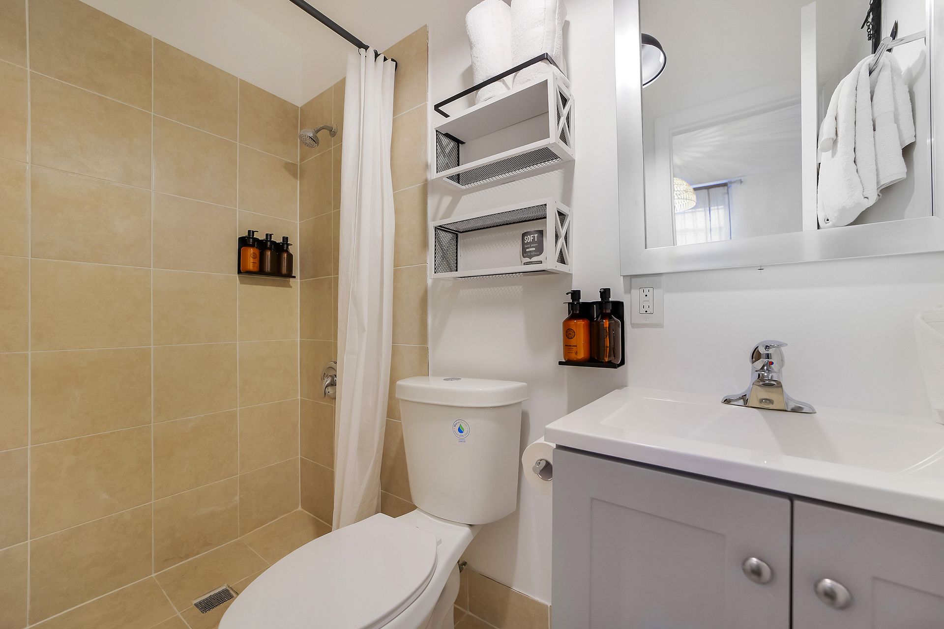 Small bathroom with beige tile, white toilet, vanity, and shower.  Shelving unit holds toiletries.