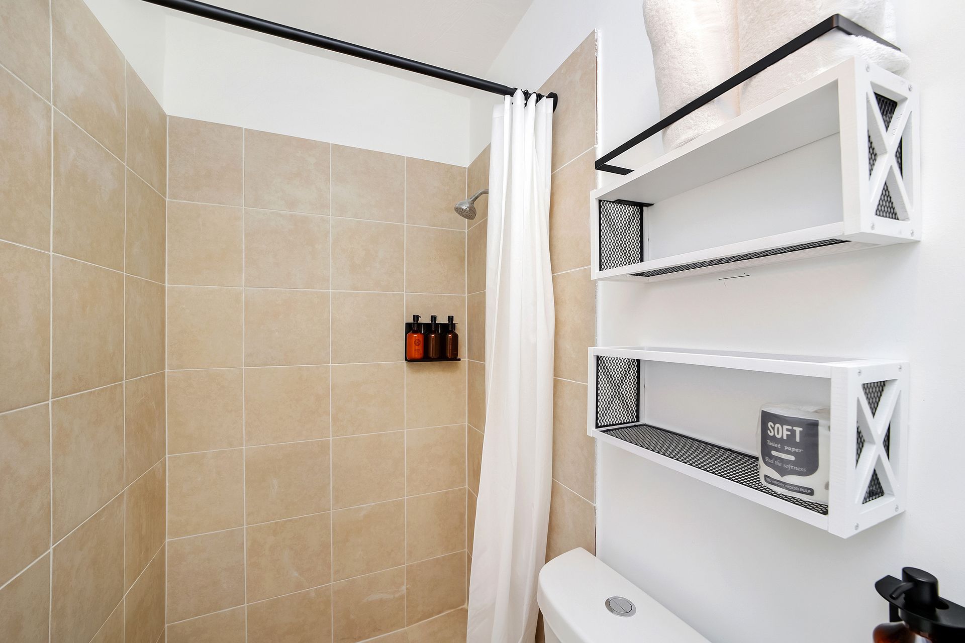 Bathroom with tan tiled shower, white curtain, shelves with towels, and a toilet.