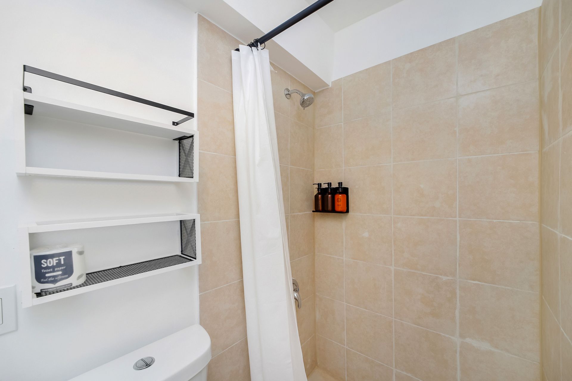 Bathroom with tiled shower and white curtain. Shelf with toilet paper and toiletries.