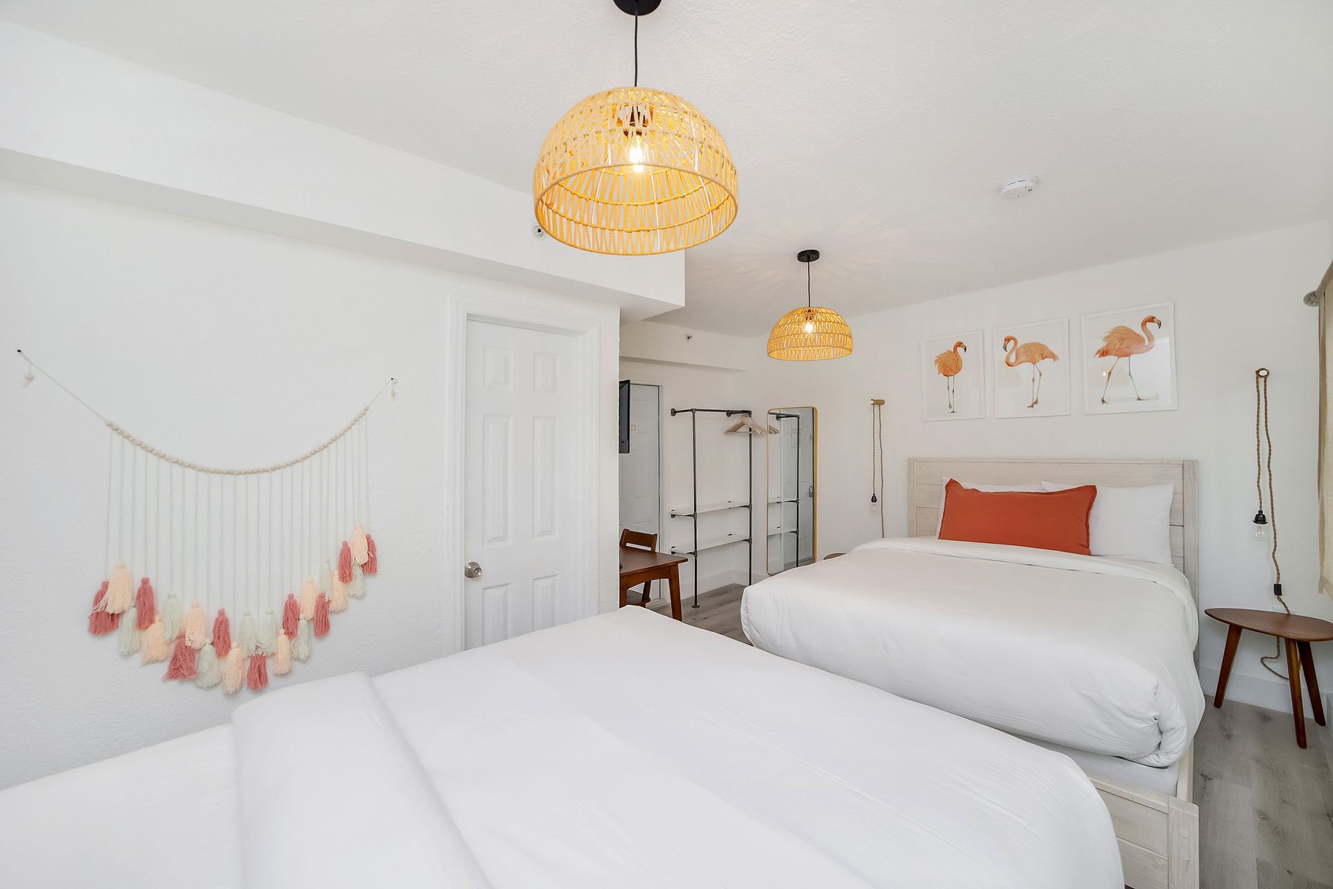 Bedroom with two beds, woven light fixtures, and flamingo art. White walls, wood accents, and cozy bedding.