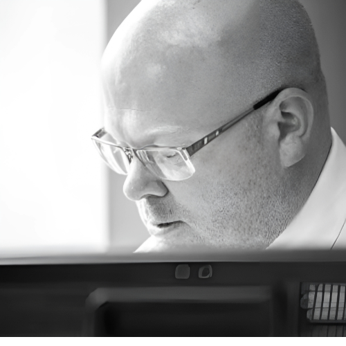 Bald man wearing glasses looking at a computer screen, focused. B&W.