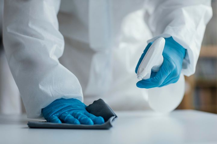 A person in a protective suit and blue gloves is cleaning a table with a cloth.