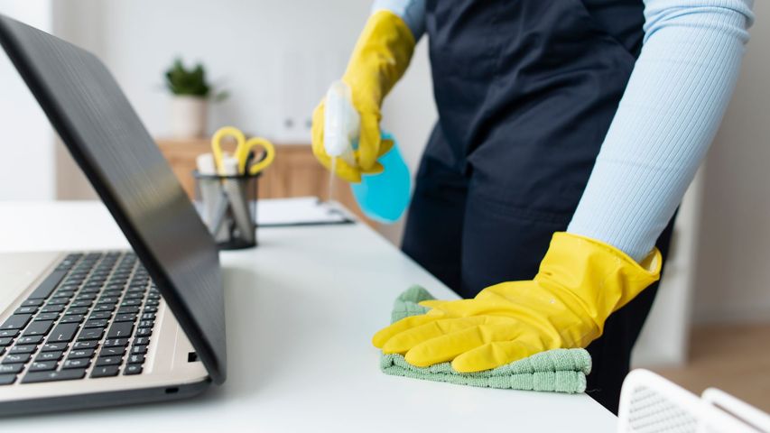 A person wearing yellow gloves is cleaning a desk in front of a laptop computer.