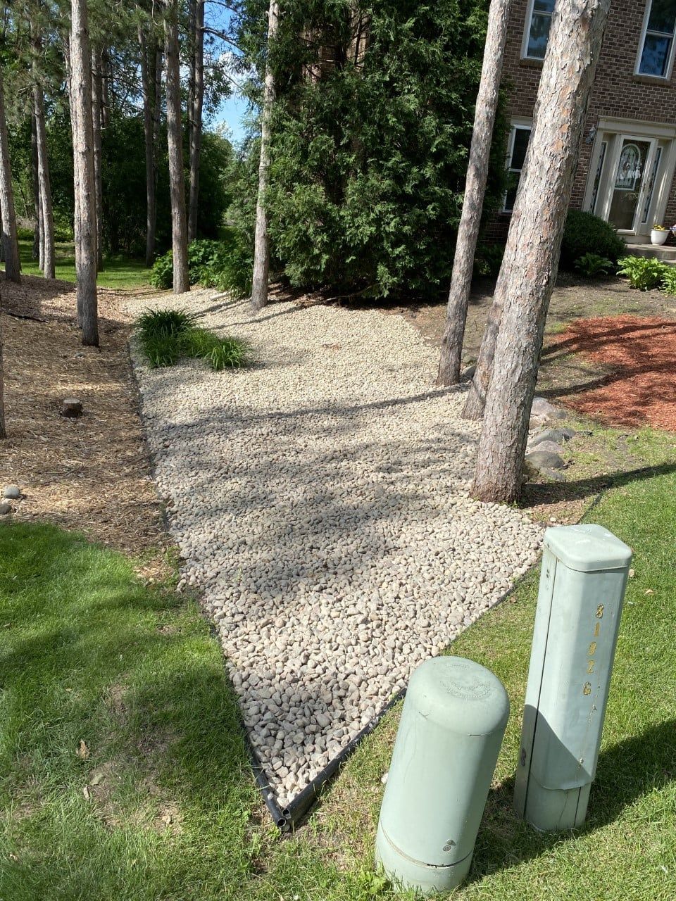 A triangular patch of landscaping stones beneath several pine trees, next to two utility pedestals and a grass lawn.
