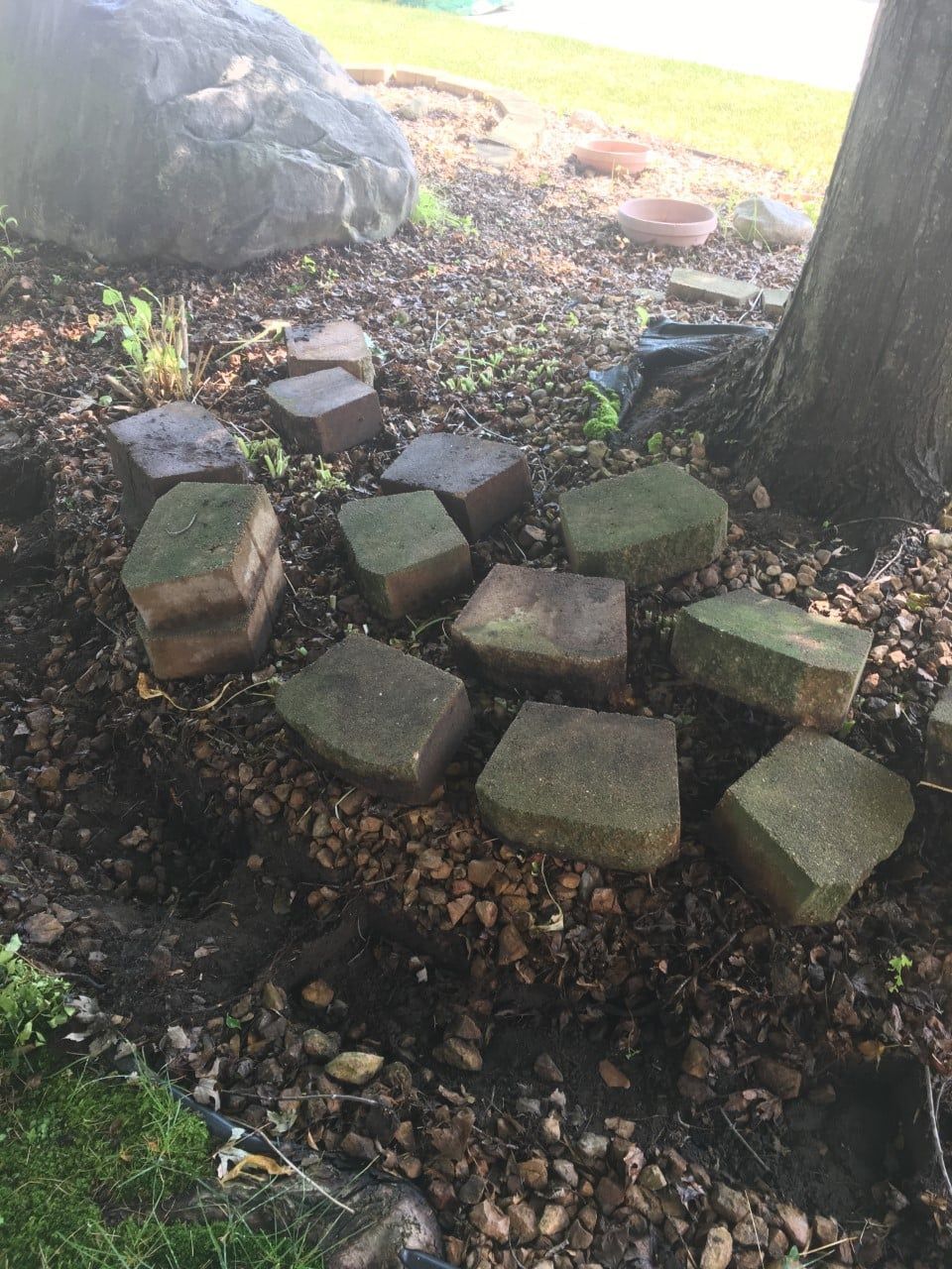 A scattering of rectangular, weathered landscape bricks resting on mulch beneath a tree near a large rock.