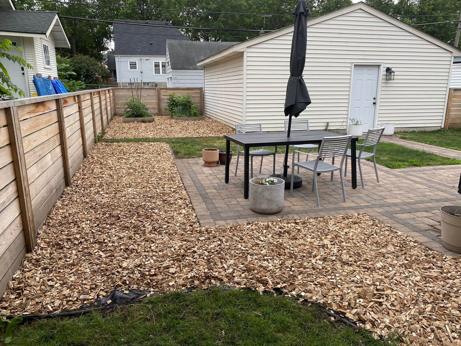 A backyard patio with a dining table and chairs, surrounded by wood-chip mulch and a wooden fence near a white garage.