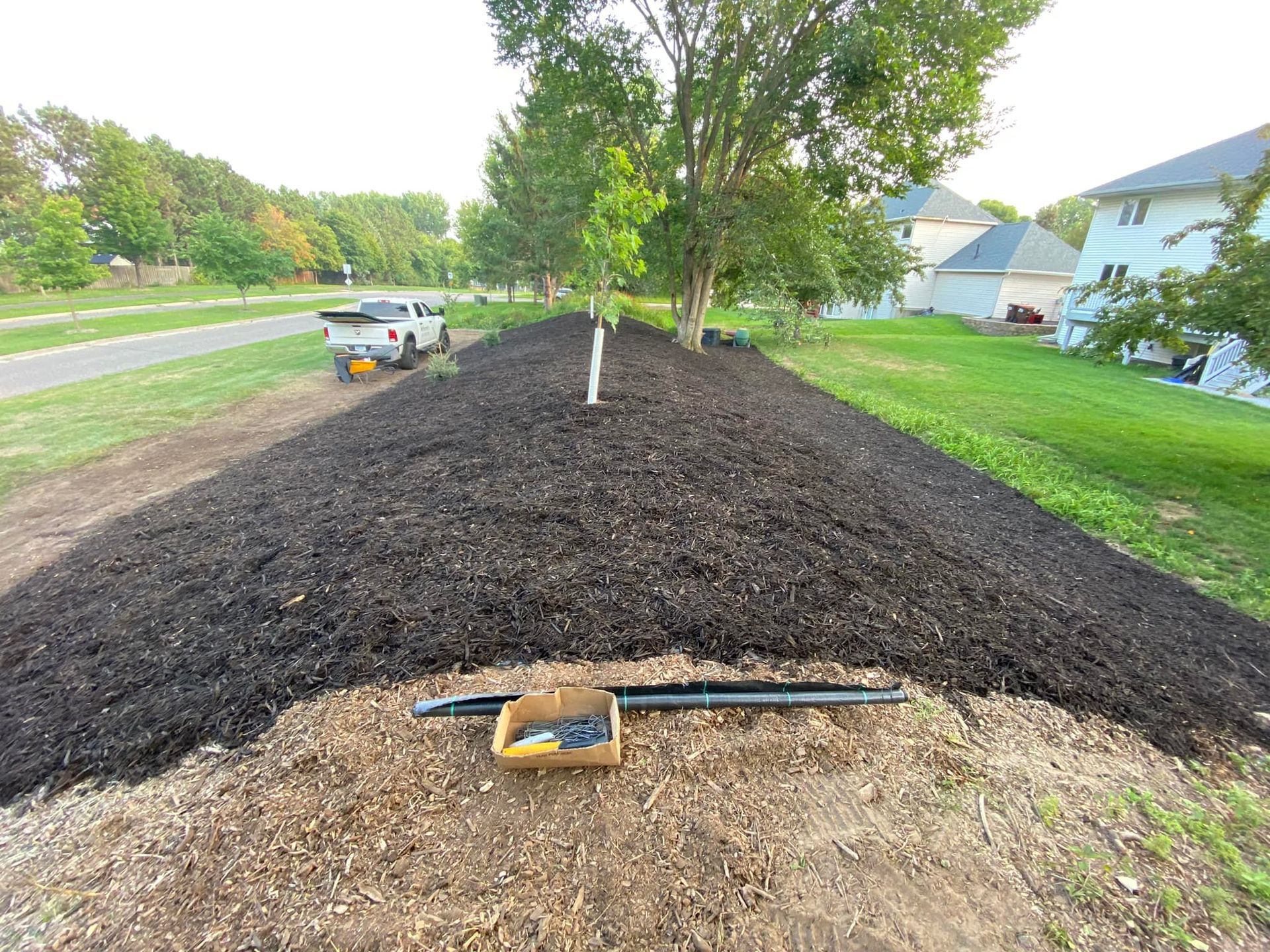 A freshly mulched garden bed sits next to a lawn and driveway, featuring a small tree and a box of irrigation supplies.