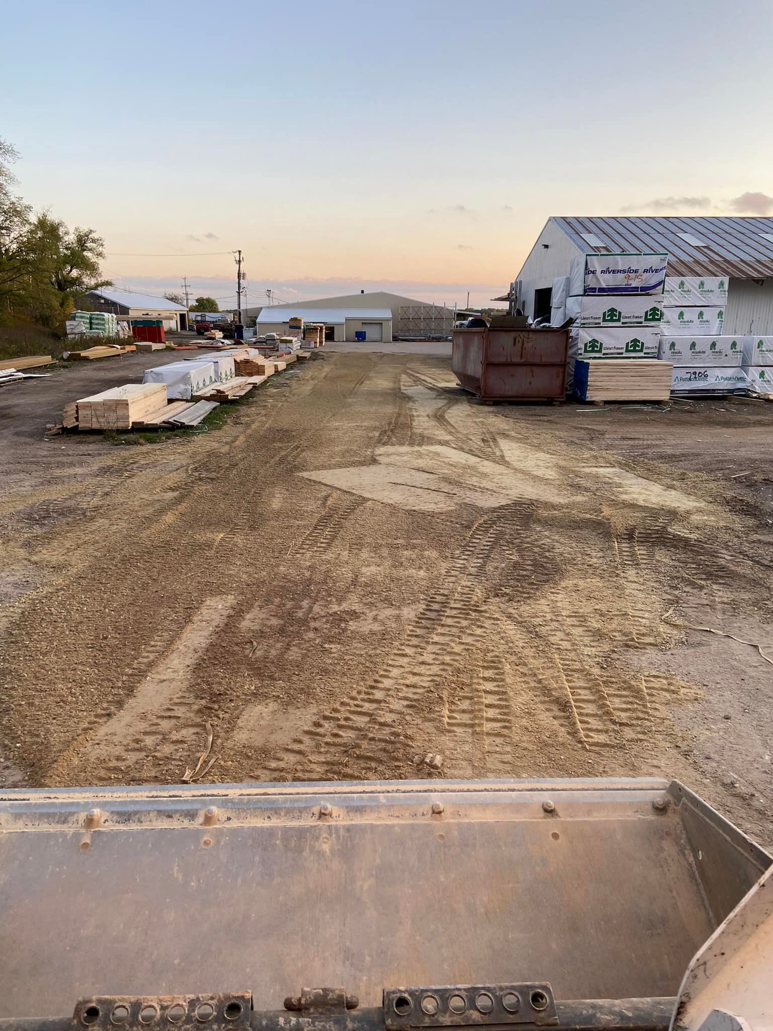 View from inside construction equipment looking down a dirt lot at a building site with piles of supplies and a dumpster.
