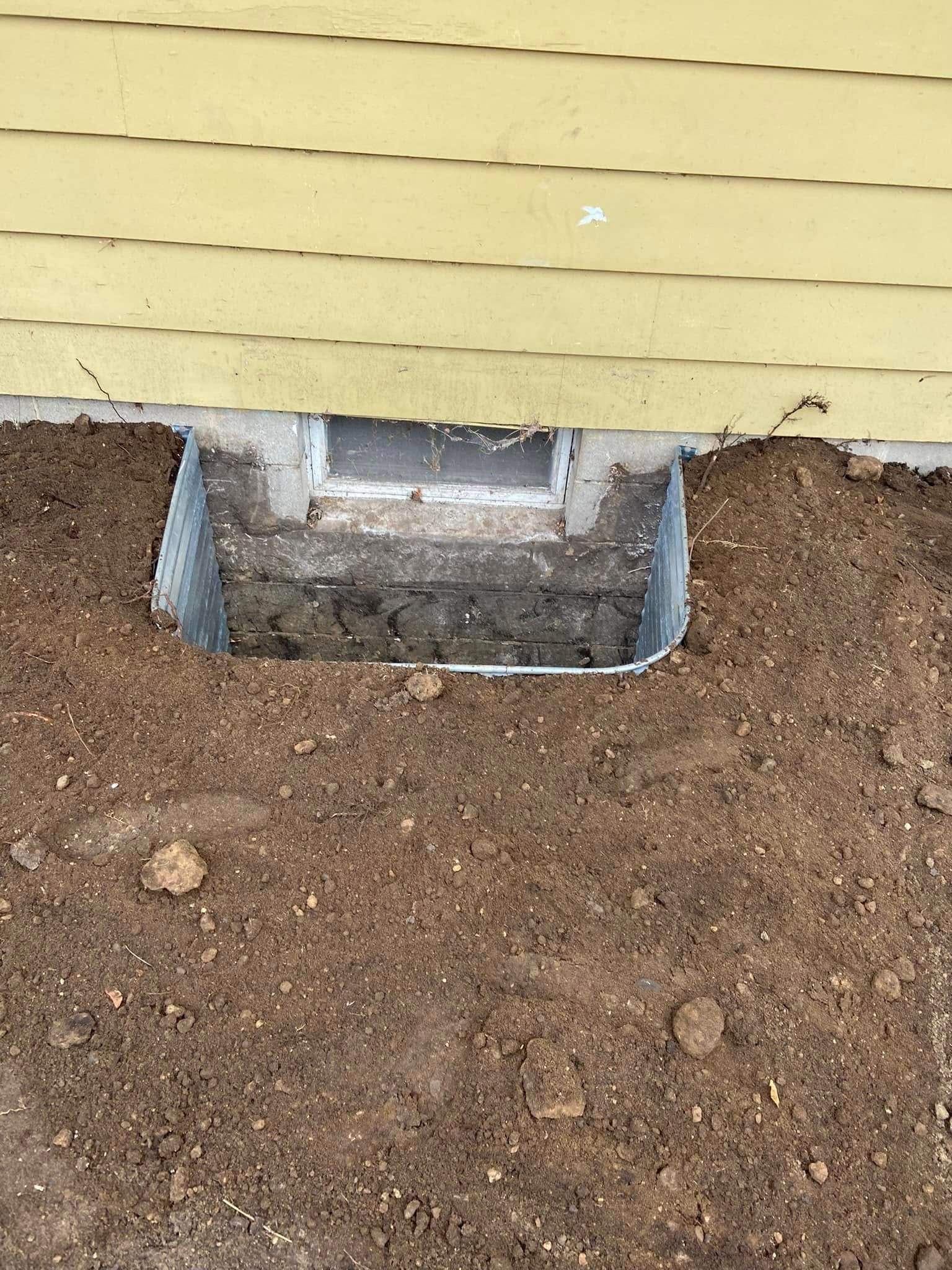 A metal window well installed against the concrete foundation of a yellow house, surrounded by bare soil.