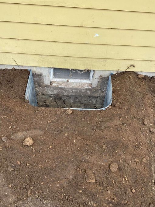A metal window well installed against the concrete foundation of a yellow house, surrounded by bare soil.