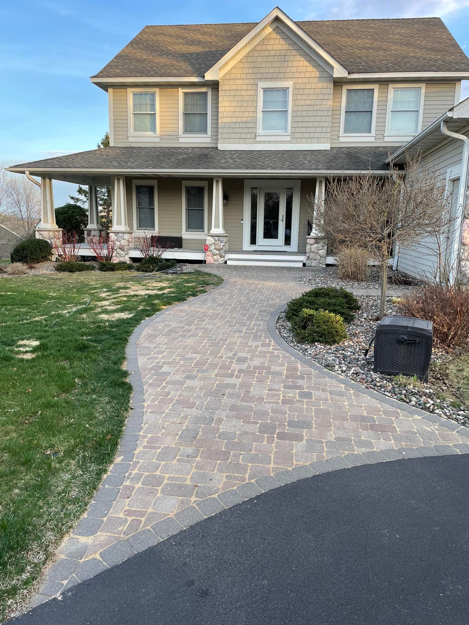 A two-story tan house with a wrap-around porch, a paved walkway, and a partial view of a driveway under a blue sky.