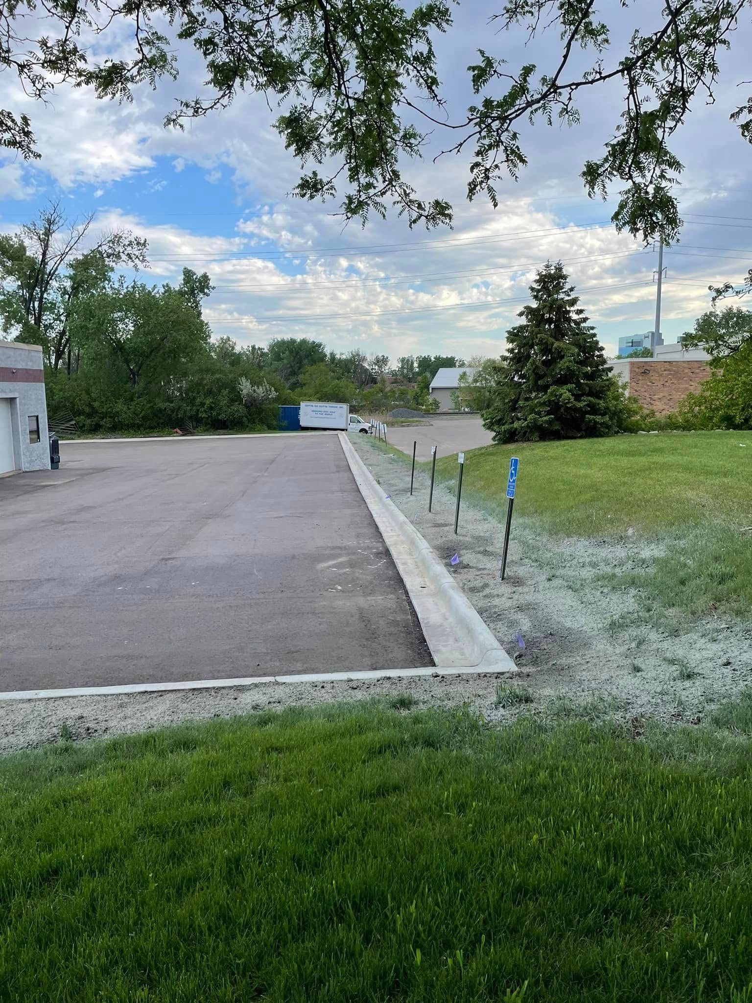An asphalt parking lot borders a grassy lawn with a line of small metal signs, under a partly cloudy sky.