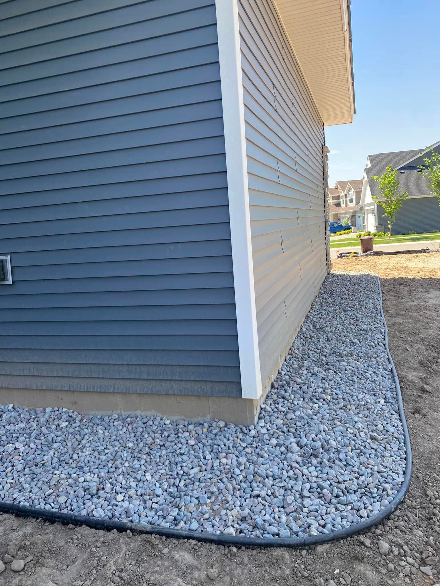 A side view of a house with dark gray horizontal siding, a tan foundation, and a curved border filled with gray gravel.