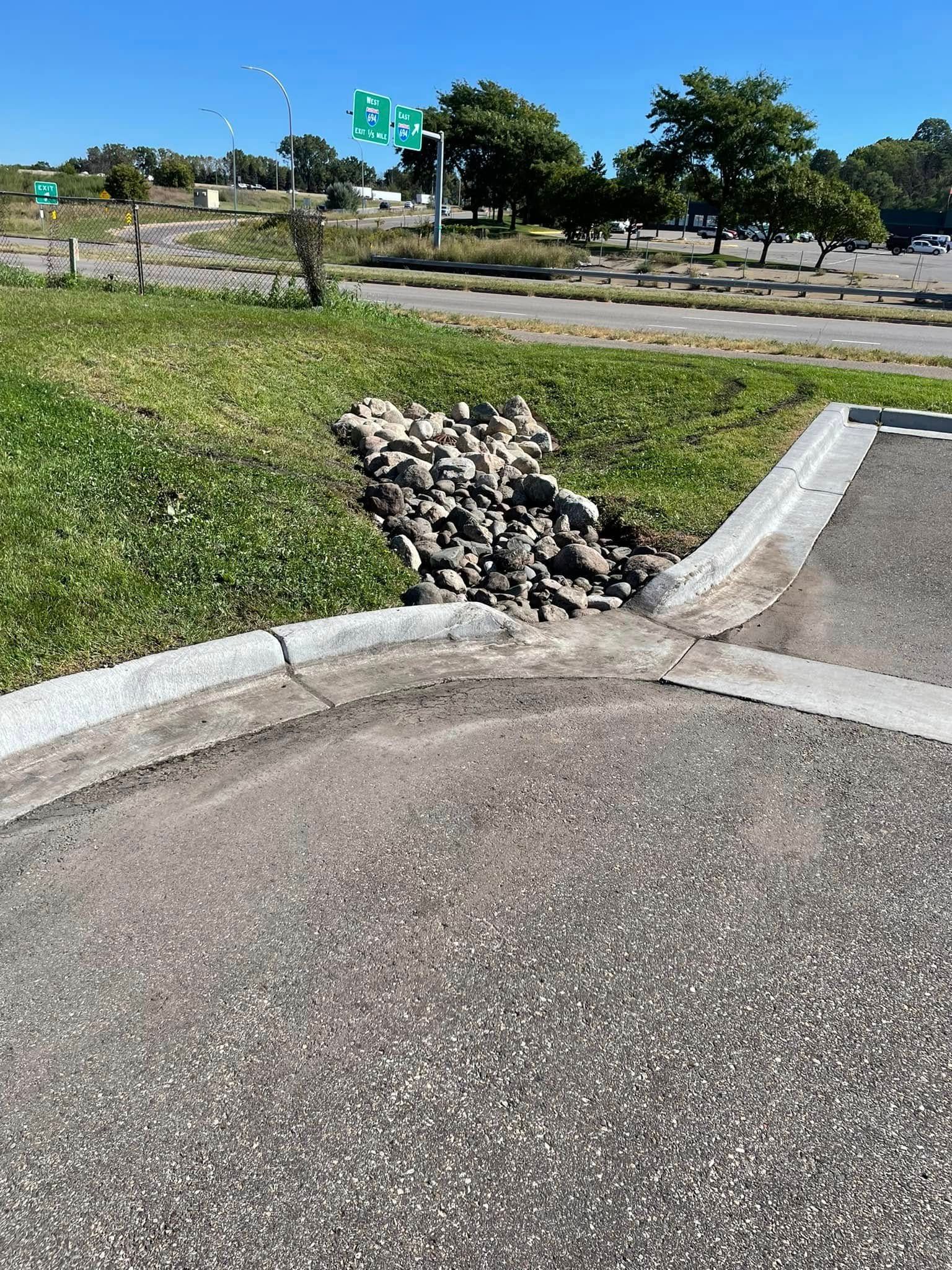 A pile of rocks in a grass verge alongside a concrete curb next to an asphalt road and a highway exit sign.