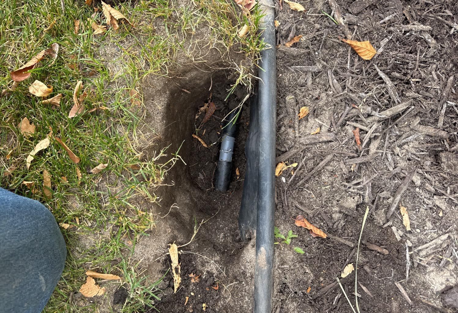 A small hole in the ground reveals a black pipe with a coupling, positioned next to a dark gray pipe in mulch and grass.