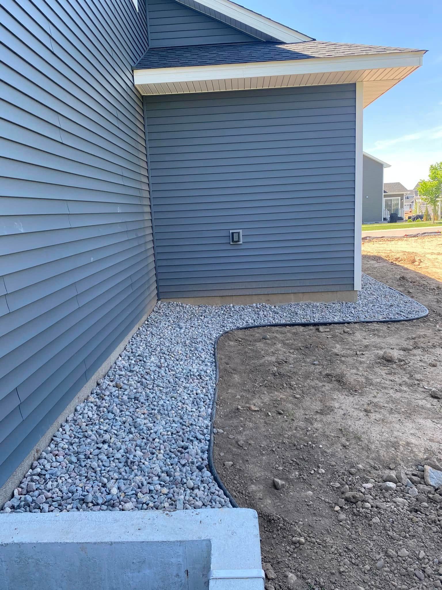 A gray-sided house corner with a freshly laid stone gravel border against the foundation and an unlandscaped dirt yard.