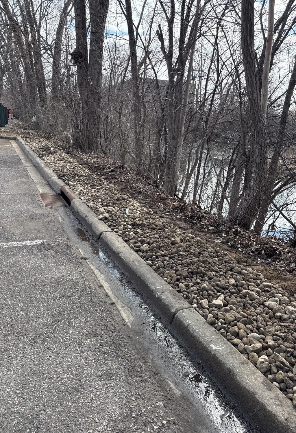 A concrete curb separates an asphalt parking lot from a gravel embankment with trees overlooking a body of water.
