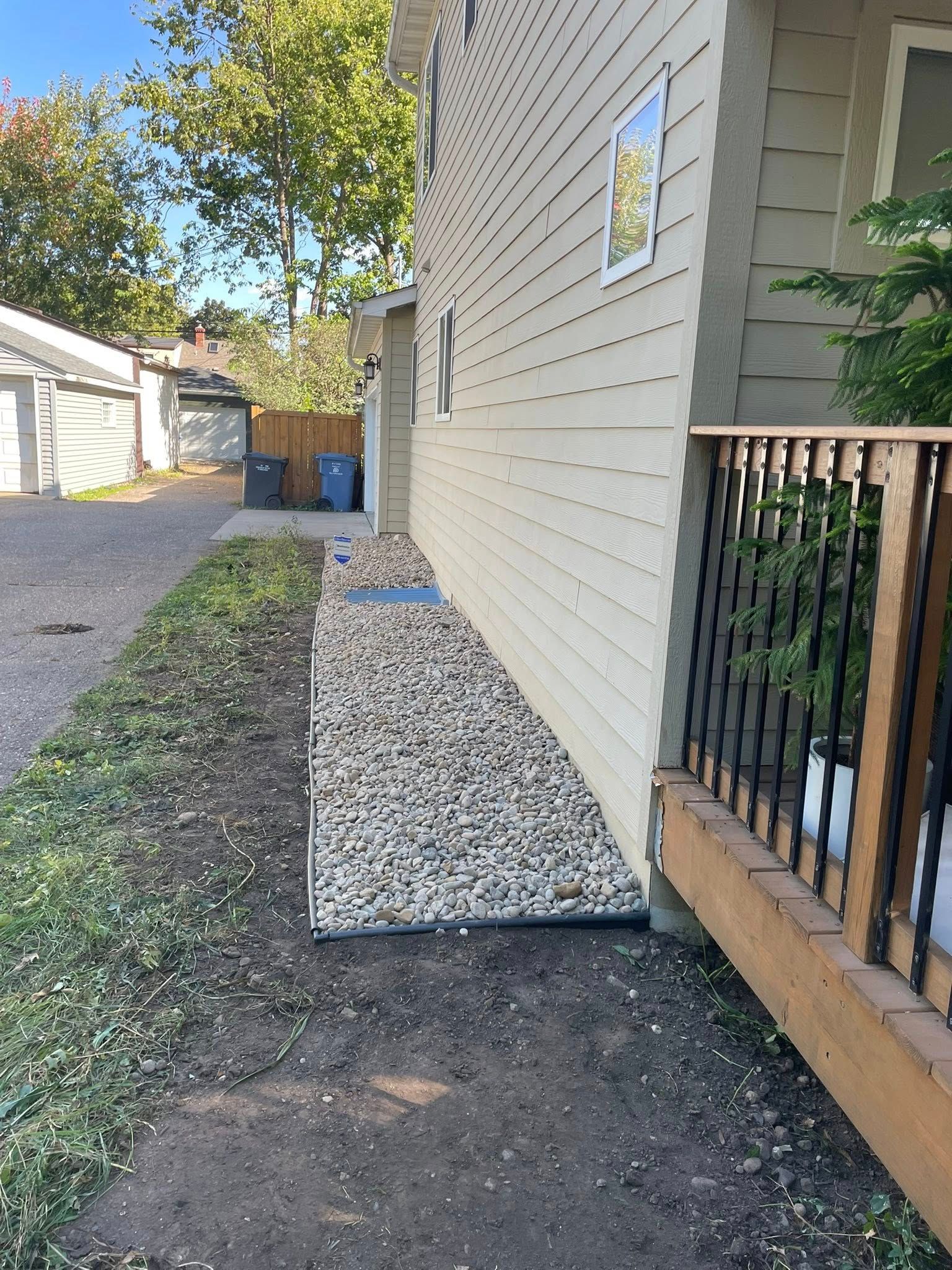 A strip of gravel mulch runs along the beige siding of a house, next to a wooden deck railing and dirt ground.