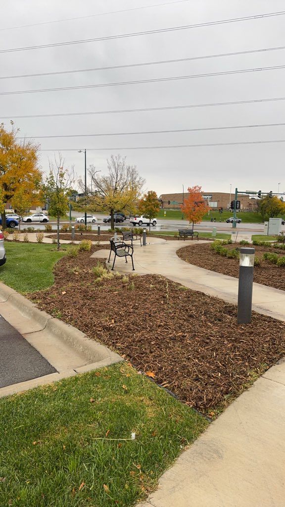 A person walks with a rollator along a paved path in a park-like area with landscaping, trees, and distant parked cars.