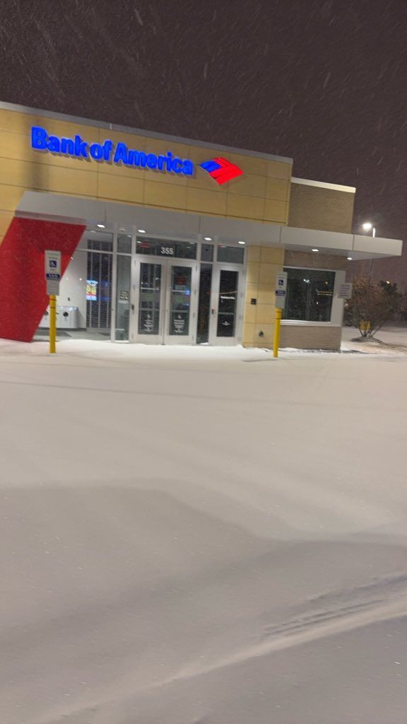 A snow-covered parking lot in front of a Bank of America branch at night during a snowfall.
