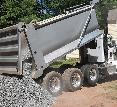 A dump truck in a residential area unloading a pile of crushed grey stone from its raised bed onto the dirt ground.