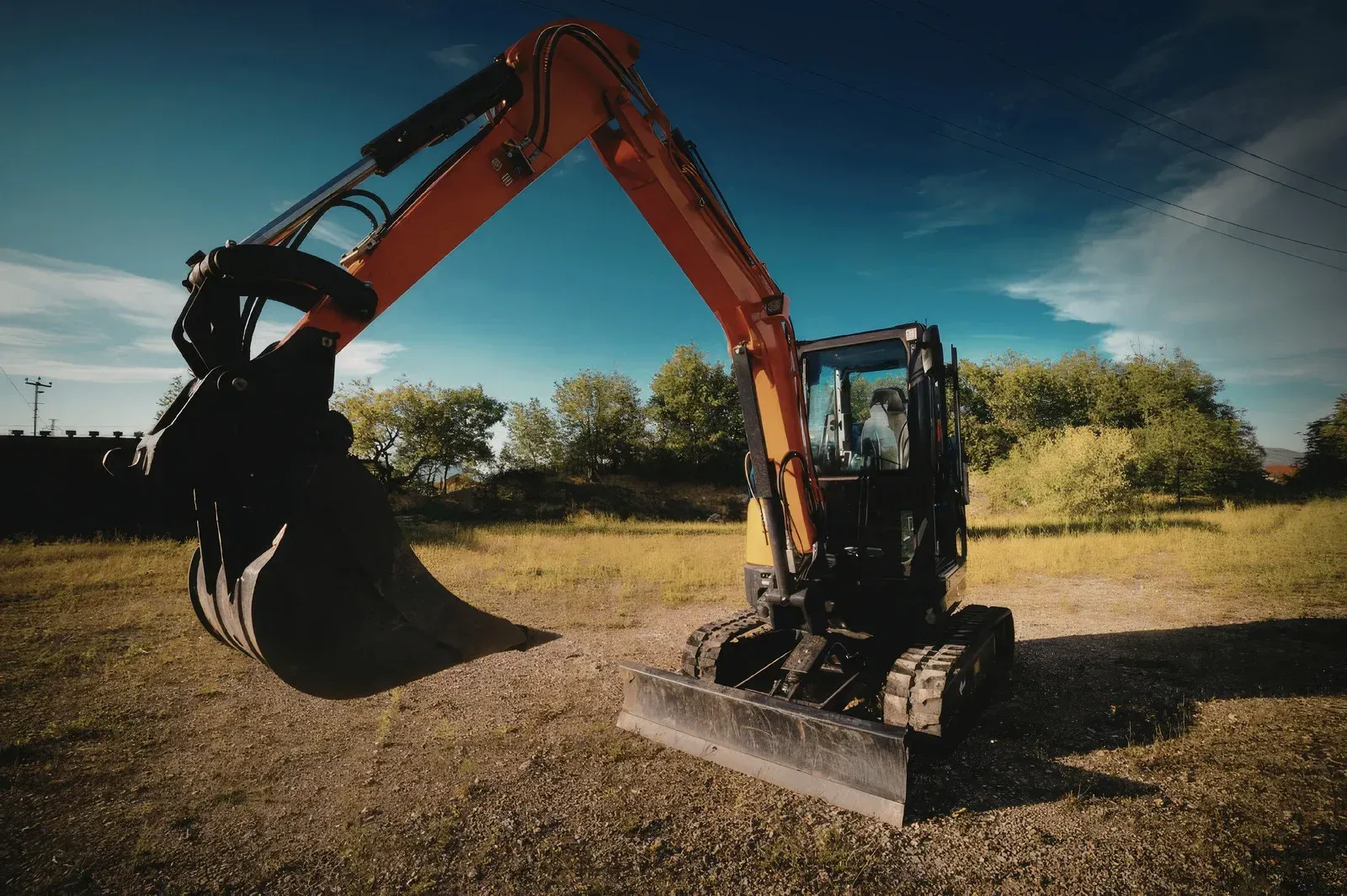 An orange mini excavator stands in a grassy field under a sunny blue sky, with its digging arm raised.